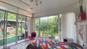 Un dormitorio moderno con ventanas amplias, un edredón colorido, una silla roja y vistas a un jardín.