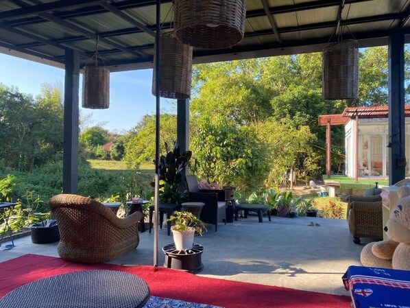 Un espacio de estar al aire libre cubierto con mobiliario de mimbre, plantas en macetas y vista a un jardín y un edificio al fondo.