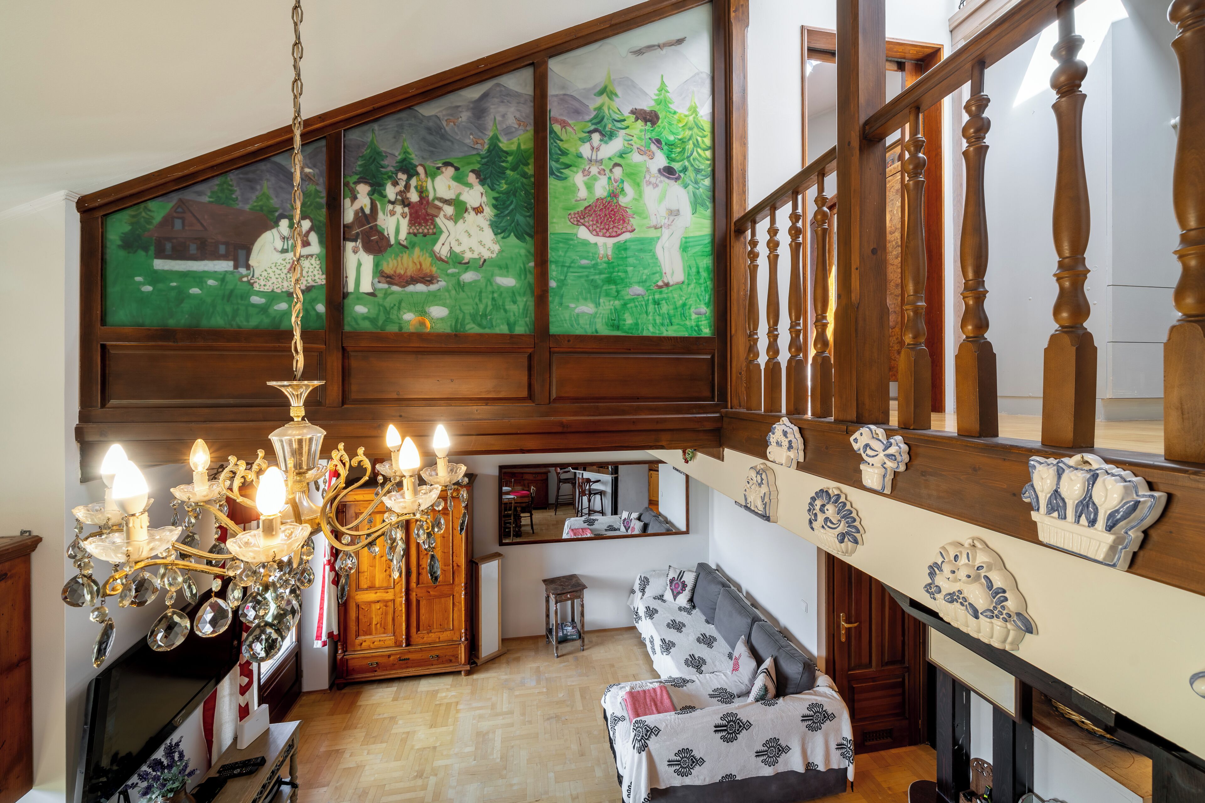 A room with a wooden staircase, a chandelier, a sofa, a mirror, and a large window with a painting.