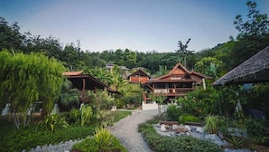 A traditional wooden building with a thatched roof, surrounded by lush greenery and a gravel path.