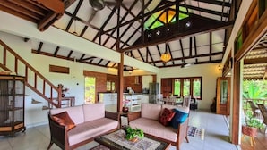 A spacious living area with a high ceiling, wooden beams, a sofa set, a coffee table, and a view of the kitchen in the background.
