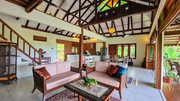 A spacious living area with a high ceiling, wooden beams, a sofa set, a coffee table, and a view of the kitchen in the background.
