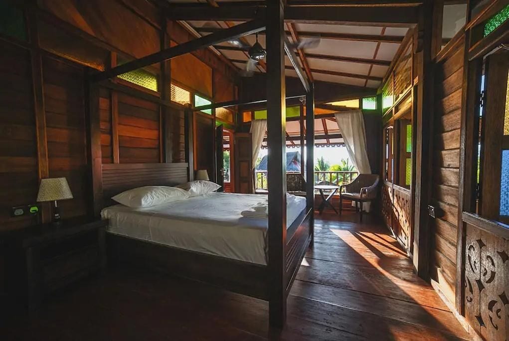 A wooden four-poster bed in a room with a balcony, a nightstand with a lamp, and a view of the outdoors.