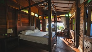 A wooden four-poster bed in a room with a balcony, a nightstand with a lamp, and a view of the outdoors.