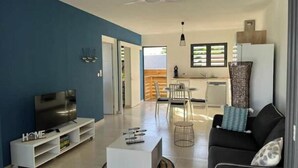 Uma sala de estar moderna com televisão, unidade de entretenimento branca e um sofá. Há uma área de jantar com cadeiras e uma mesa, e uma cozinha com uma janela.