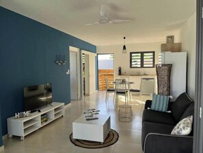 A modern living room with a TV, white entertainment unit, and a sofa. There's a dining area with chairs and a table, and a kitchen with a window. - Villa TiSable - Your relaxing break just a stone's throw from the beach (Saint-Paul)