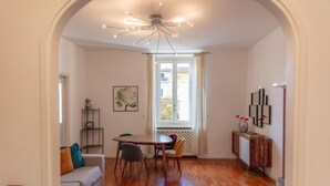 A dining area with a round table, chairs, a sofa, a decorative ceiling light, and a window with curtains.