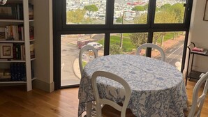 En matplats med ett bord och stolar, en bokhylla och ett stort fönster med utsikt över en småbåtshamn och stadens silhuett.