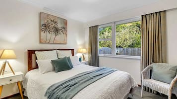 A bedroom with a bed, a chair, a window with curtains, a nightstand, and a lamp.