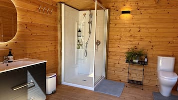 A wooden cabin bathroom with a shower, toilet, sink, and a small shelf with toiletries.
