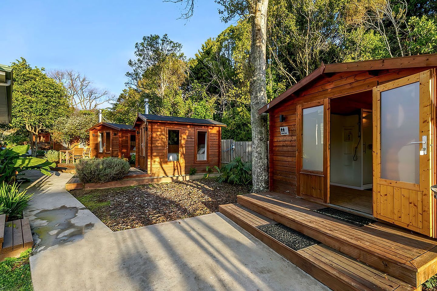 A wooden cabin with a concrete pathway leading to the entrance, surrounded by greenery.