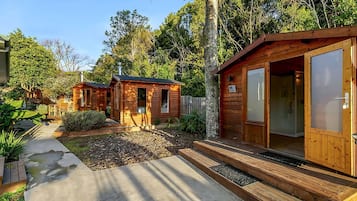 A wooden cabin with a concrete pathway leading to the entrance, surrounded by greenery.
