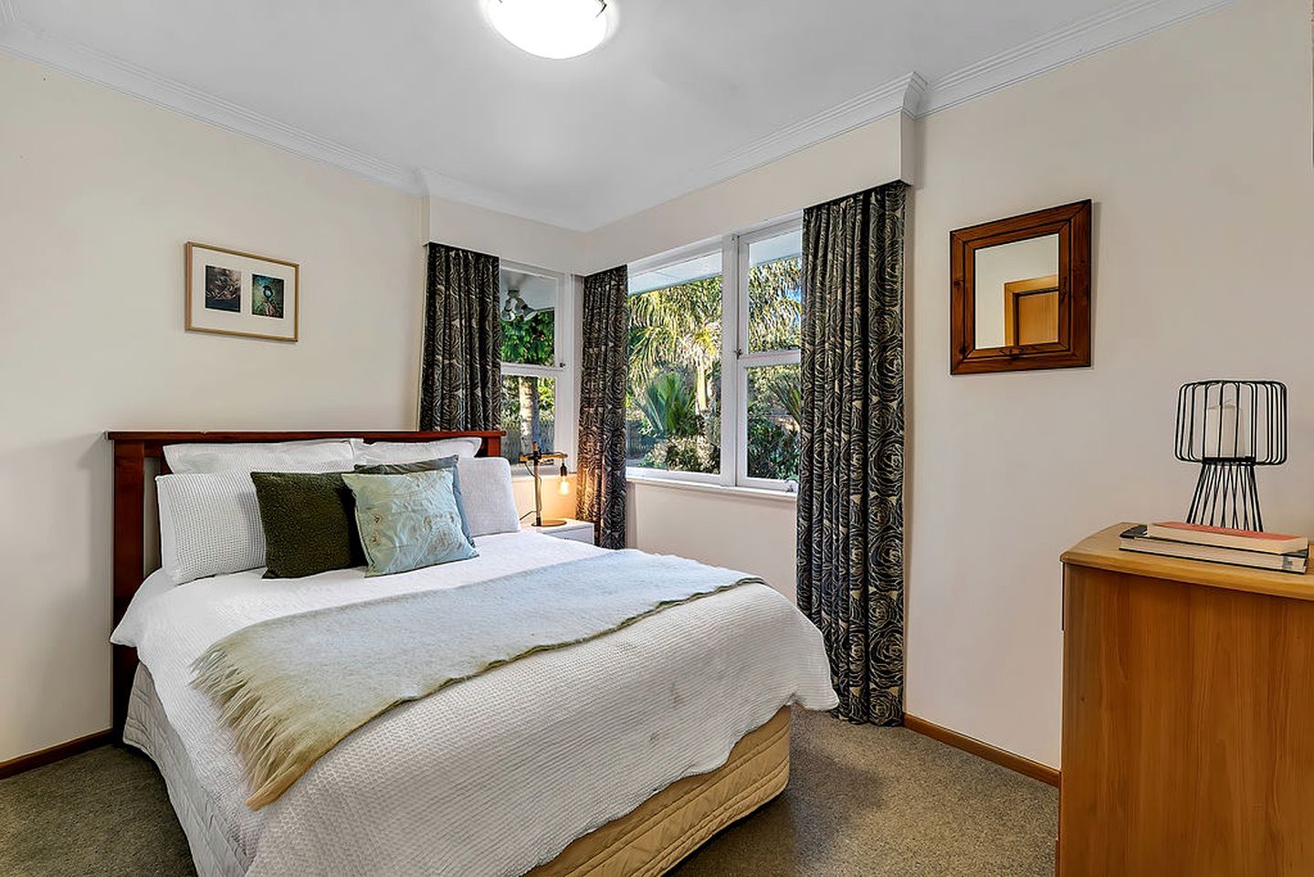 A bedroom with a bed, a bedside table, a lamp, a window with curtains, and a framed picture on the wall.