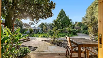 A wooden deck with a bench, surrounded by a garden with various plants and a house in the background.
