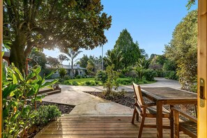 Een houten terras met een bankje, omgeven door een tuin met diverse planten en een huis op de achtergrond.