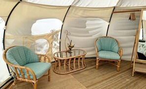 A tent with two wicker chairs and a small table, a hanging light fixture, and a bed frame against the wall.
