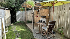 Une terrasse en bois avec une table et des chaises, un pot de fleurs suspendu et un petit coin cuisine extérieur.