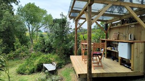 A wooden deck with a table and chairs, a kitchen area with a sink and cabinets, and a view of the surrounding greenery. - Sunny Vineyard-View Glamping with Outdoor Kitchen near Oneroa, Waiheke Island (Isla Waiheke)