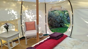 A tent with a bed, a chair, a small table, and a view of greenery outside.