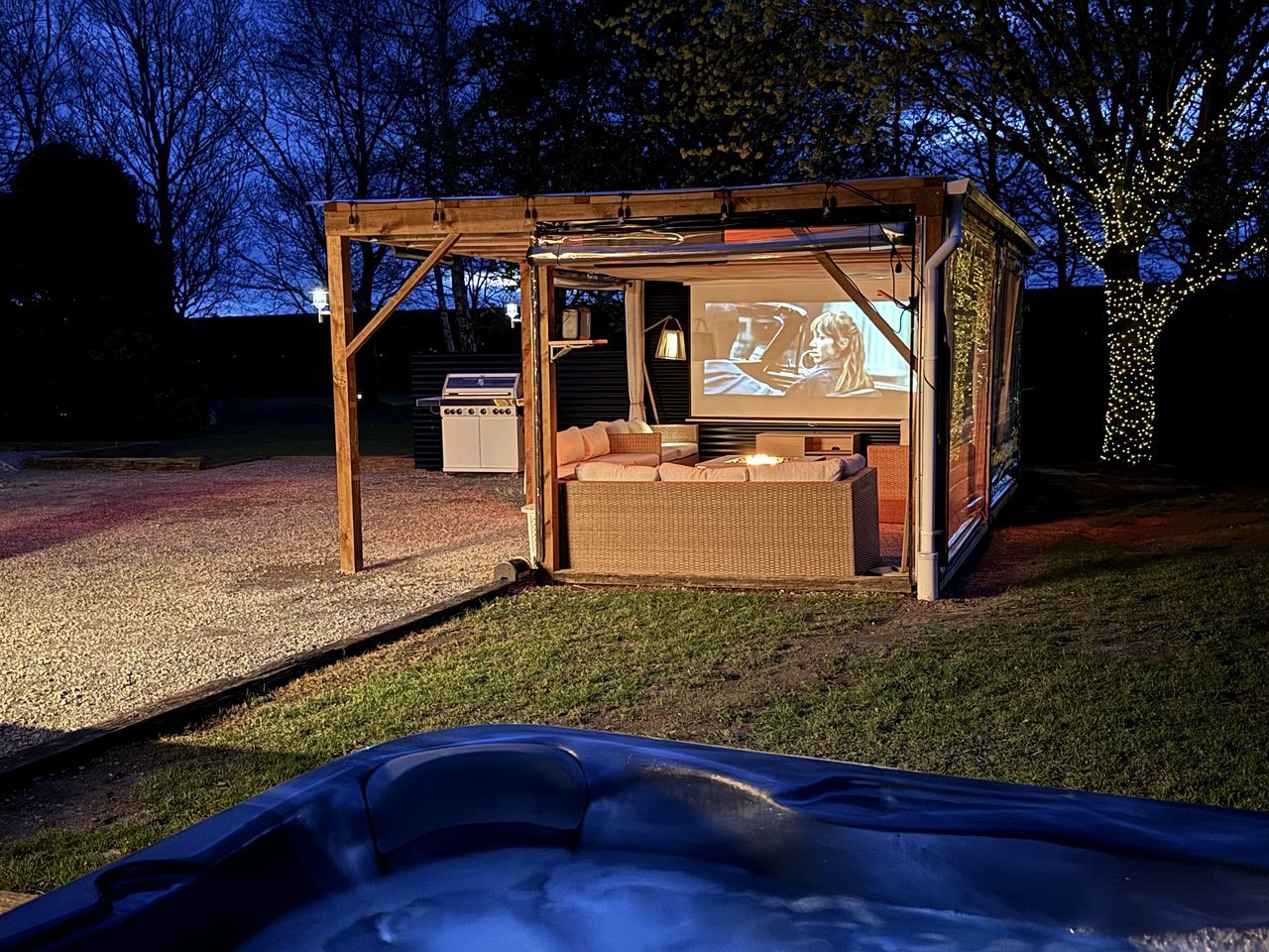 A wooden structure with a screen displaying a movie, a sofa, and a hot tub in the foreground.