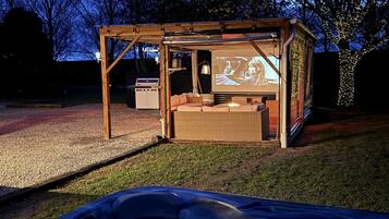A wooden structure with a screen displaying a movie, a sofa, and a hot tub in the foreground.