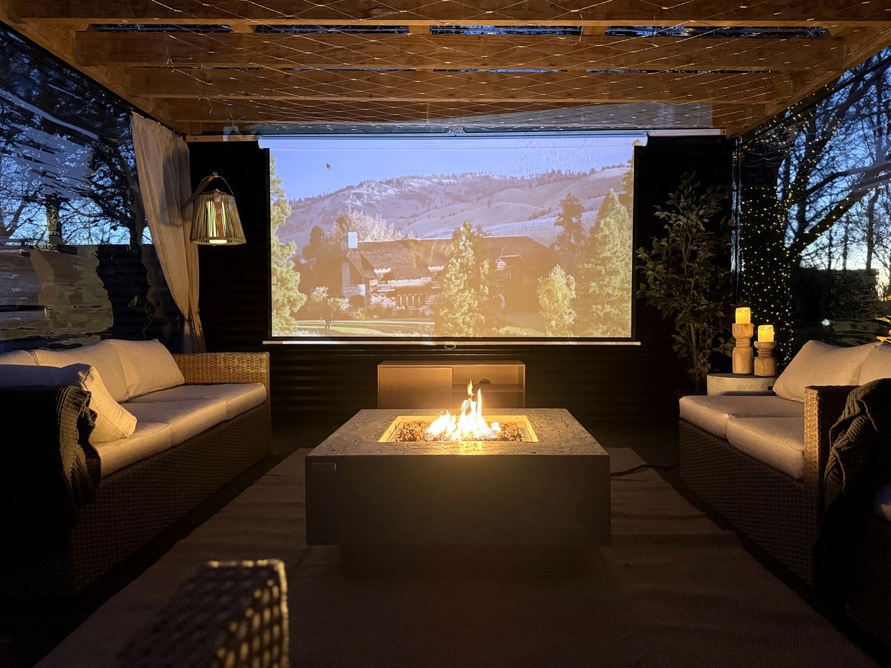 A cozy outdoor seating area with a fire pit, comfortable sofas, and a large screen displaying a scenic view.