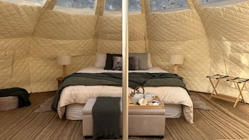 A tented room with a bed, bedside lamps, a tray with tea and pastries, and a wooden bench.