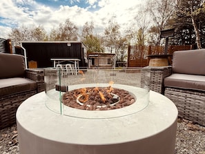 A circular outdoor seating area with a fire pit in the center, surrounded by wicker furniture and a pergola structure in the background.