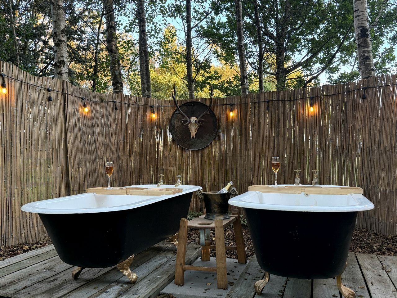 Two black bathtubs on a wooden deck with a wooden fence and a deer skull decoration in the background.