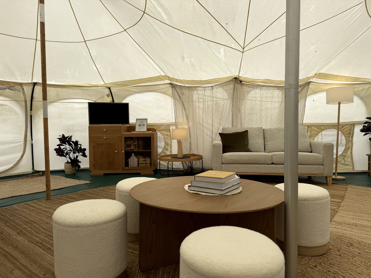 A tented accommodation with a round wooden table, two ottomans, a sofa, a cabinet, a lamp, and a plant.