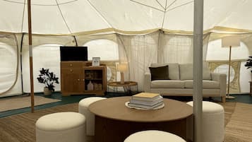 A tented accommodation with a round wooden table, two ottomans, a sofa, a cabinet, a lamp, and a plant.