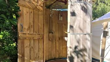 Une douche extérieure en bois, dotée d’une porte, d’un banc en bois et d’un tronc d’arbre, installée dans un jardin.