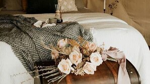 Un dormitorio con un cabecero capitonado, una cama con edredón blanco, un baúl decorativo y un ramo de flores.