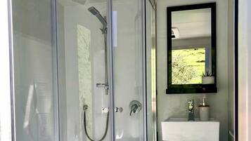 A glass-enclosed shower with a handheld showerhead, a white sink with a mirror above it, and a wooden bench.