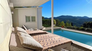 Eine Liege am Pool mit Blick auf Berge, einem kleinen Fenster und einer weißen Außenwand.