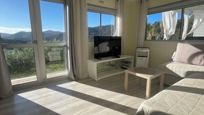Ein Zimmer mit Fernseher auf einem weißen Schrank, einem kleinen Tisch, einer Couch und großen Fenstern mit Vorhängen.