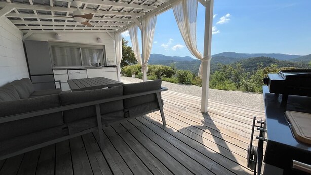 Ein überdachter Außenbereich mit einer Sitzecke, einem Sofa, einem Tisch, einem Grill und Blick auf Berge.