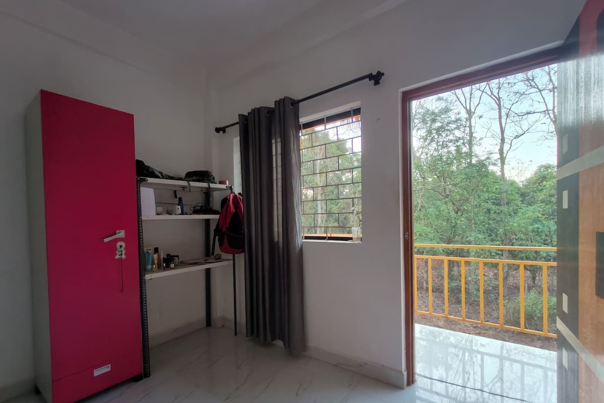 A room with a red wardrobe, a desk with shelves, a window with curtains, and a balcony with a yellow railing.