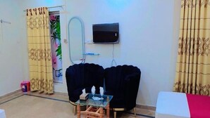 A living room with two dark sofas, a glass coffee table, a TV mounted on the wall, and a small table with a tissue box and glasses. - Hotel Shelton One (Multan)