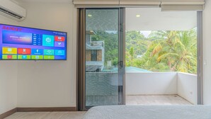Une chambre d’hôtel moderne dotée d’un grand téléviseur à écran plat affichant diverses applications, d’une porte-fenêtre coulissante donnant sur un balcon avec vue sur la verdure, et d’un lit avec un matelas à texture particulière.