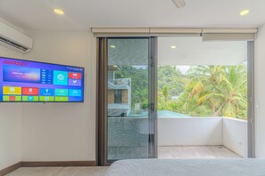 Ein modernes Hotelzimmer mit einem großen Flachbildfernseher, der verschiedene Apps anzeigt, einer Schiebetür zu einem Balkon mit Blick ins Grüne und einem Bett mit einer strukturierten Matratze.