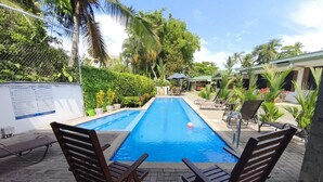 Une piscine rectangulaire à l’eau d’un bleu limpide, entourée d’une terrasse en briques. On y voit deux chaises longues en bois, face à la piscine, et un petit parasol est visible au fond.