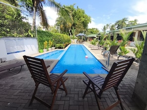 Ein rechteckiger Swimmingpool mit klar blauem Wasser, umgeben von einer Ziegelterrasse. Zwei Holzliegen sind zum Pool hin ausgerichtet, und im Hintergrund ist eine kleine Sonnenschirmkonstruktion zu sehen.