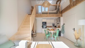 Un espace de vie de style loft avec un escalier en bois, un coin repas avec des chaises vertes, une cuisine équipée d’appareils modernes et un lit en mezzanine doté d’une tête de lit en osier.