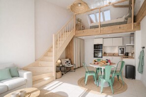 En modern lägenhet i loftstil med en trätrappa, en matplats med ett runt bord och stolar, ett litet kök och ett vardagsrum med en soffa.