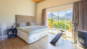 Une chambre à coucher comprenant un lit, un ventilateur sur pied, un radiateur, un banc et une vue sur une montagne, visible à travers une grande fenêtre.