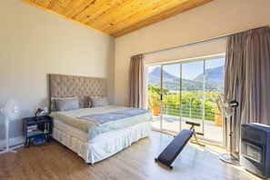 A bedroom with a bed, a standing fan, a heater, a bench, and a view of a mountain through a large window. - Greenview Villa -holiday home (Cape Town)