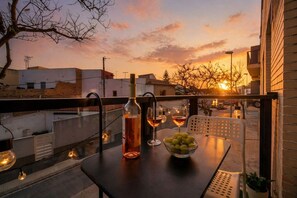 A balcony with a table set for two, featuring a bottle and glasses, and a bowl of grapes.