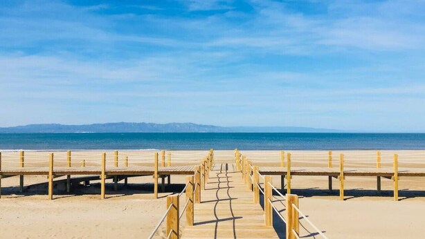 Ein hölzerner Steg führt zu einem Strand mit blauem Himmel und fernen Bergen.