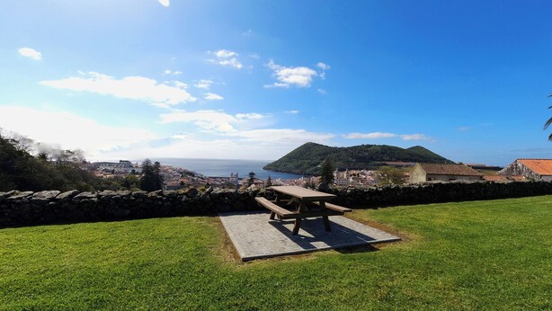 Un área con césped, una mesa de picnic, un muro de piedra y vistas a un pueblo costero con edificios y colinas al fondo.